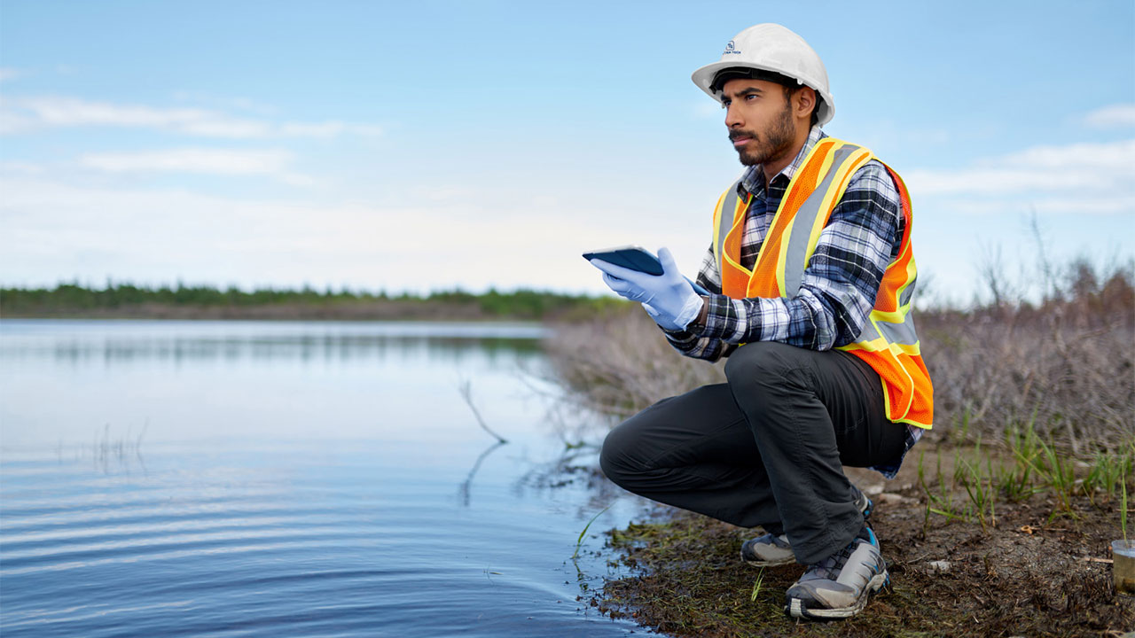A person crouched near a riverbed while holding a tablet, looking over the surrounding environment during inspection