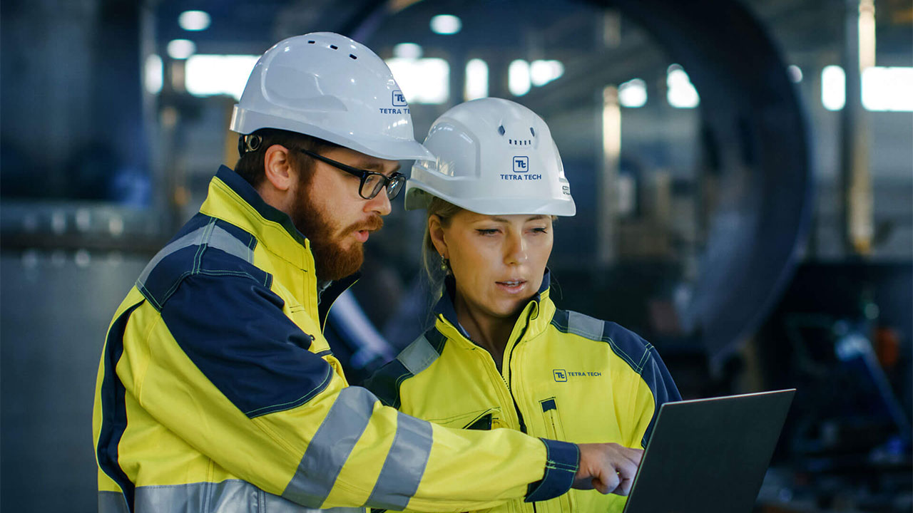 Two workers in construction equipment looking at a laptop while in a warehouse
