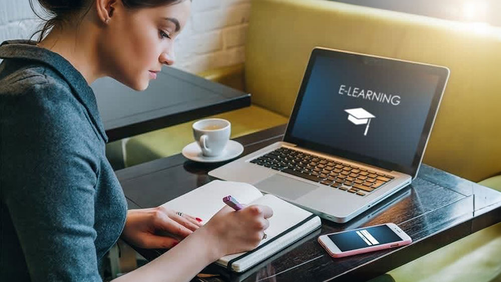 A person with an open laptop that reads "E-Learning" while taking notes in a journal